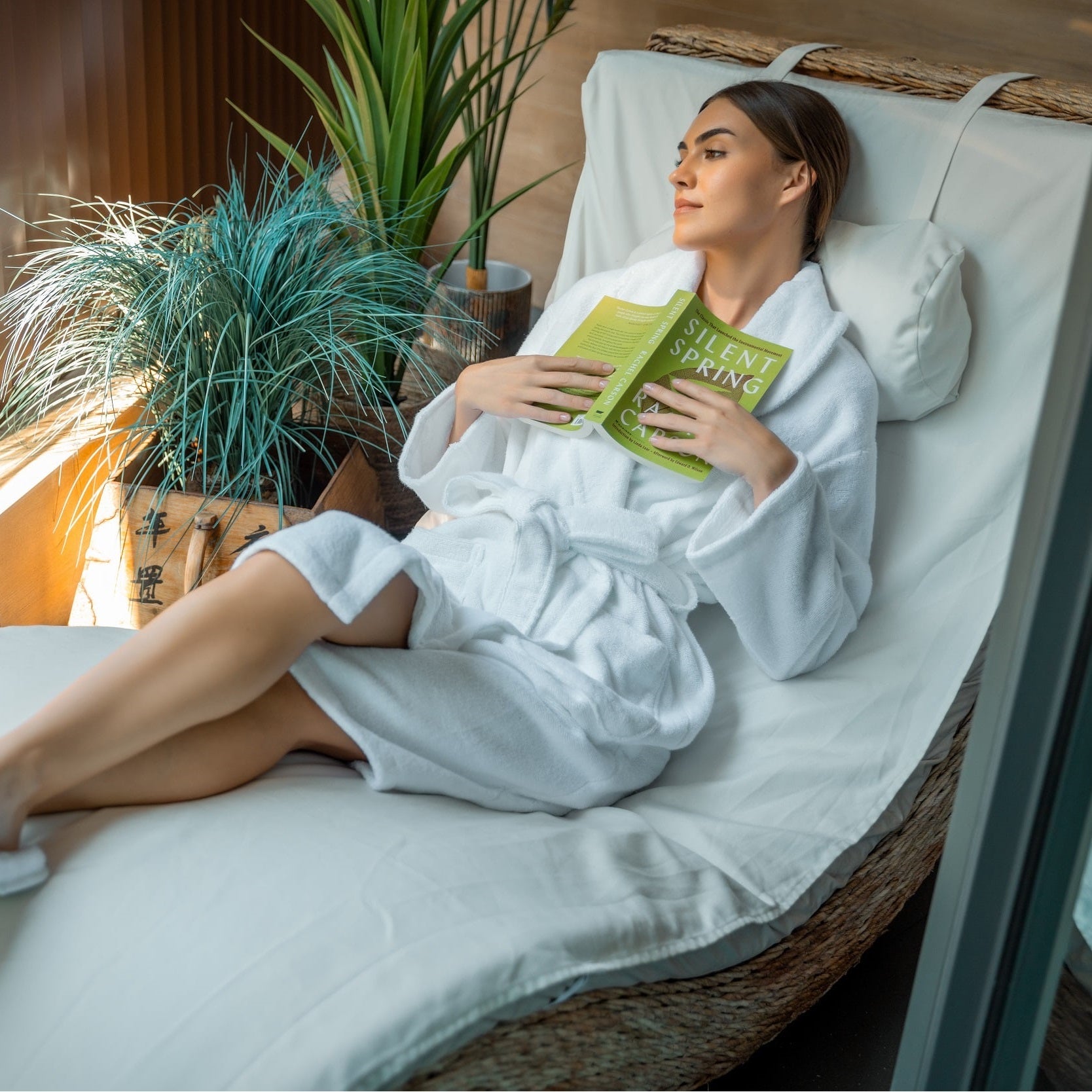 A woman in a dye-free, chlorine-free robe is lying on a sustainable wicker lounge reading Silent Spring by Rachel Carson.