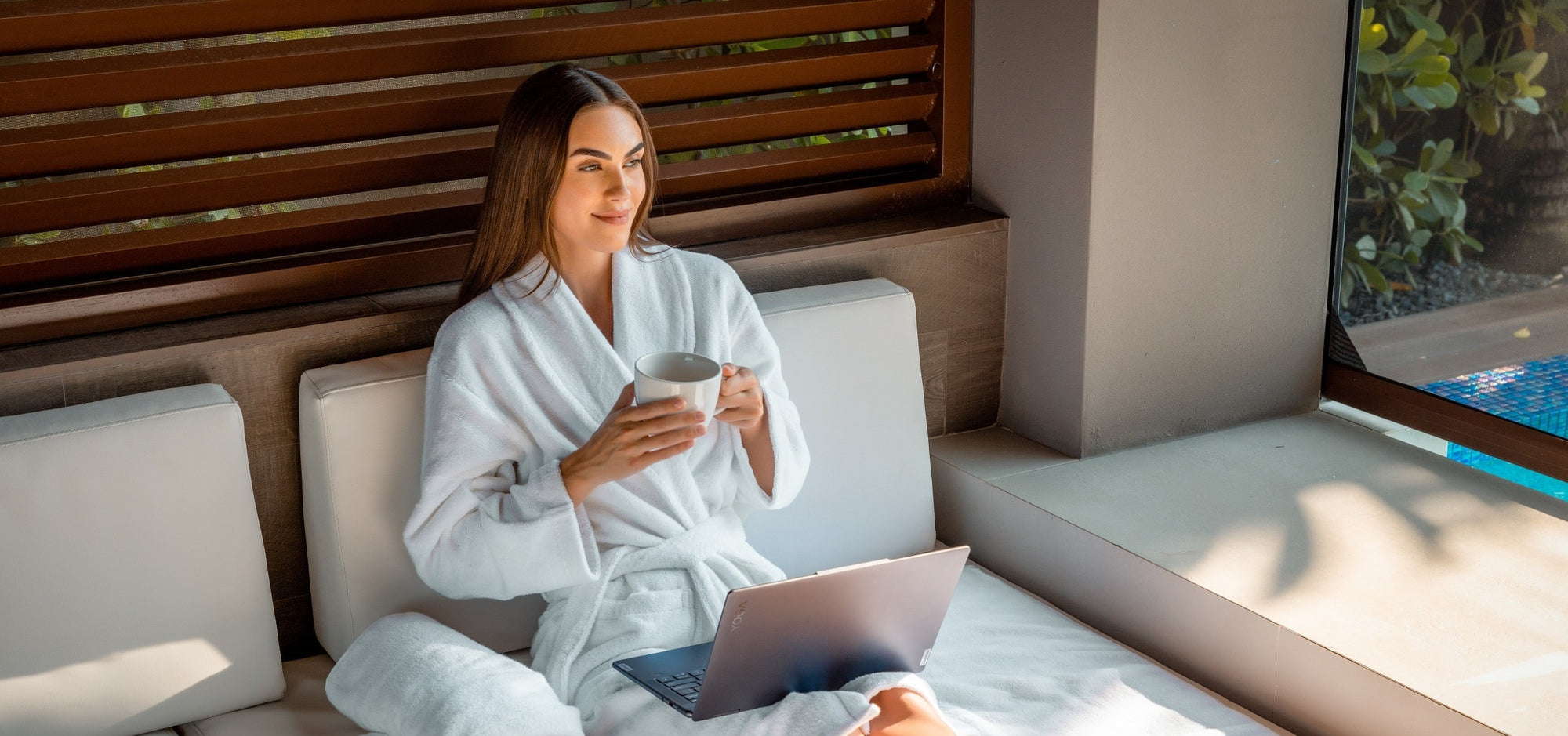 A woman lounges in an organic cotton terry robe and on organic cotton terry towels as she works on her laptop and sips her coffee.