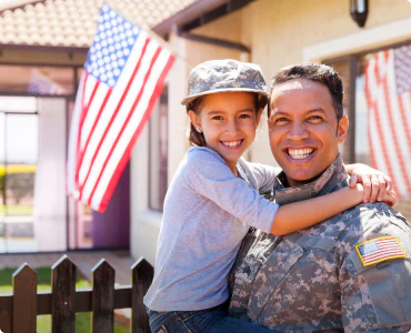 A military father holds his cute daughter as both are smiling next to an American flag, happy with their well-deserved military family discount.