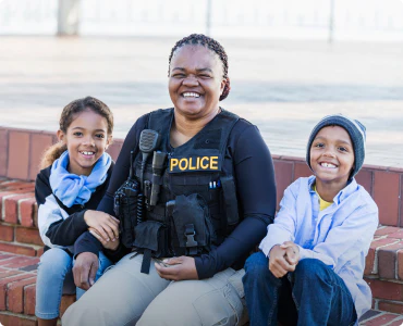 An African-American female police officer is smiling with two smiling children on her sides, happy that her family receives generous police discounts.